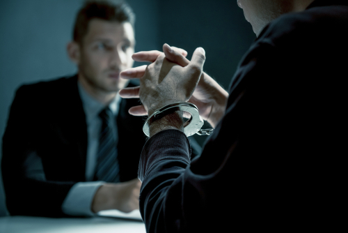 A person in handcuffs sits across from a lawyer in a formal interview room, appearing to discuss their case.