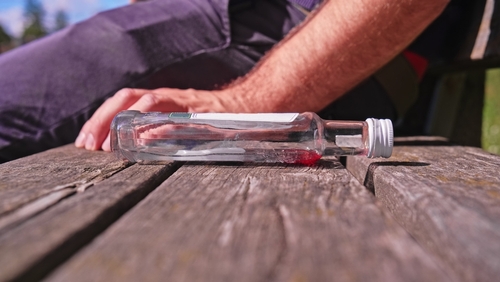 Empty liquor bottle lying on a wooden surface with a blurred image of a person passed out in the background.