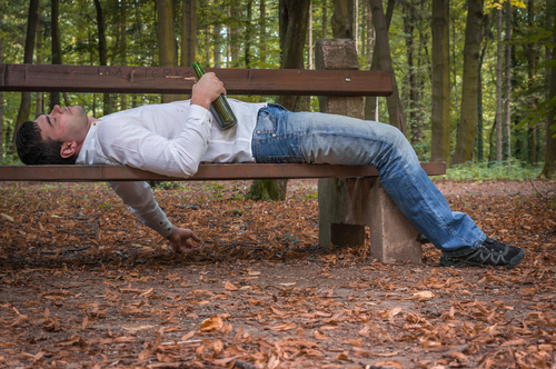 Man lying on a park bench in the woods holding a beer bottle, appearing intoxicated.