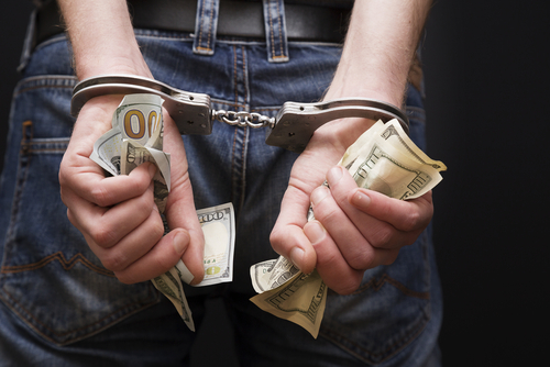 A close-up of a person's hands in handcuffs, holding multiple $100 bills, symbolizing financial theft or white-collar crime.