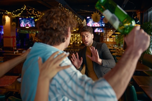 Man in a bar raises a glass bottle in a threatening manner during a heated altercation, while another man backs away in fear.