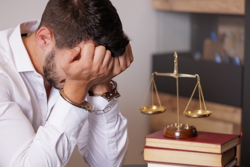 Arrested man in a white shirt sits with his head in his hands beside legal books and a justice scale, symbolizing legal stress and consequences.