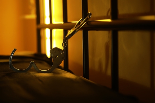 Open handcuffs hanging from a prison cell bar with dim lighting, representing incarceration and the criminal justice system.