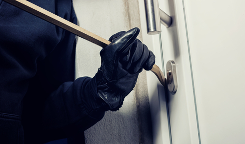 Close-up of a person wearing black gloves using a crowbar to break into a door, representing an attempted burglary or forced entry.