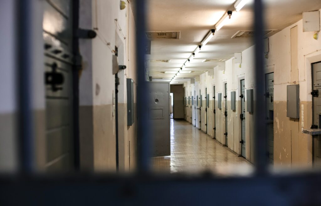 View through prison bars of a dimly lit hallway lined with closed metal cell doors in a correctional facility.