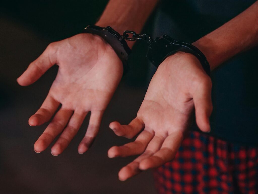 Close-up of a person’s hands in handcuffs, palms facing up, wearing a dark shirt and red checkered pants.