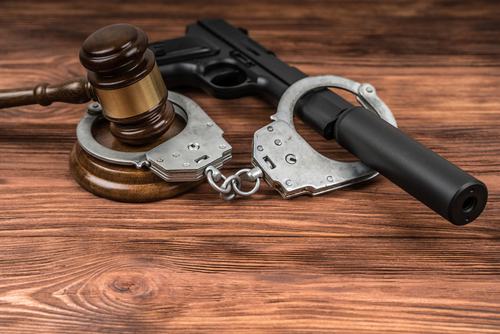 A wooden judge’s gavel, a pair of metal handcuffs, and a black handgun with a suppressor are arranged on a wooden surface, symbolizing criminal justice and law enforcement themes.