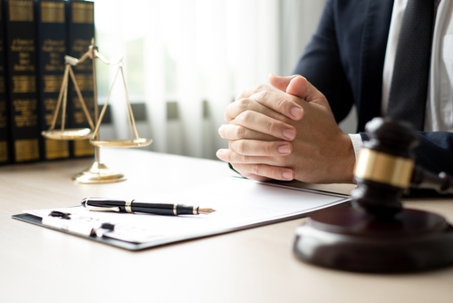 Man in formal attire sits at a legal desk with folded hands, surrounded by a gavel, justice scales, law books, and a document with a pen.