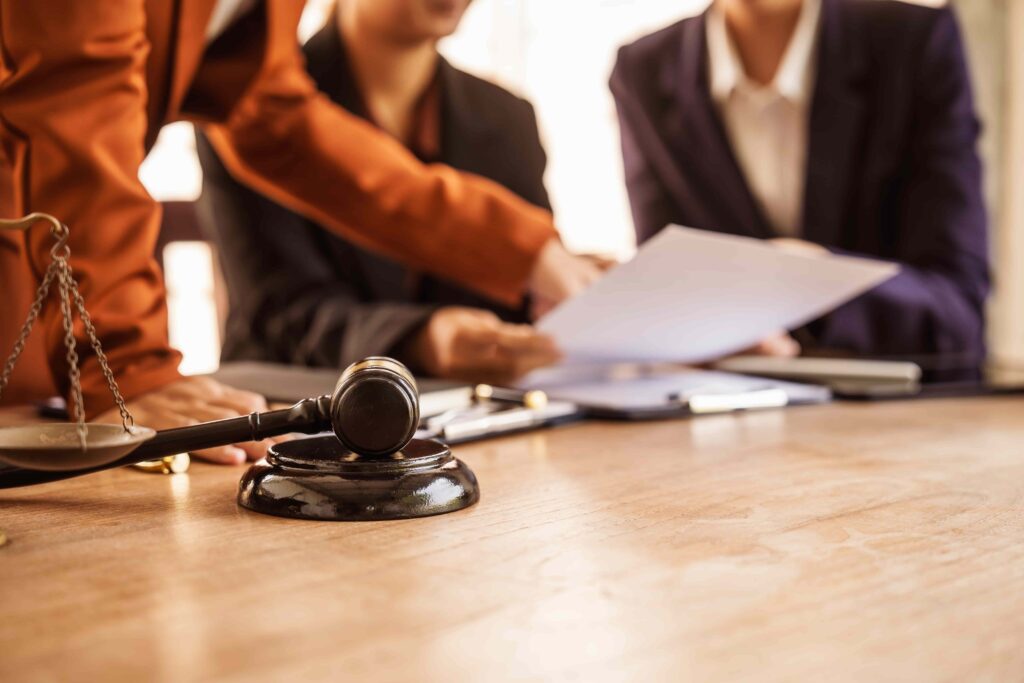 Judge&rsquo;s gavel on a courtroom table as legal professionals review documents during a court proceeding.